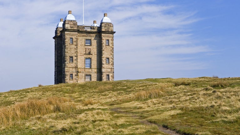 A view of the Cage, an Elizabethan hunting tower, from the grassy hillside path leading up to it, framed by blue skies.